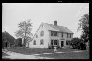 18th c. saltbox home, with woman in front yard and barn behind.